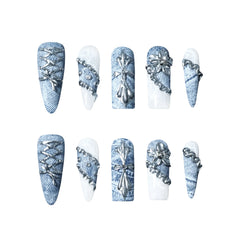 Blue denim-inspired stiletto-shaped press-on nails featuring white marble accents, metallic lace-up designs, and floral appliqués.