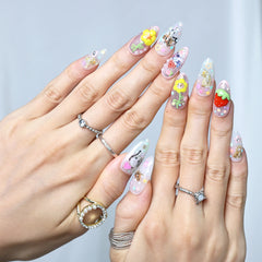 Hand model showing pink almond nails with 3D cartoon faces, strawberry charms, and floral details