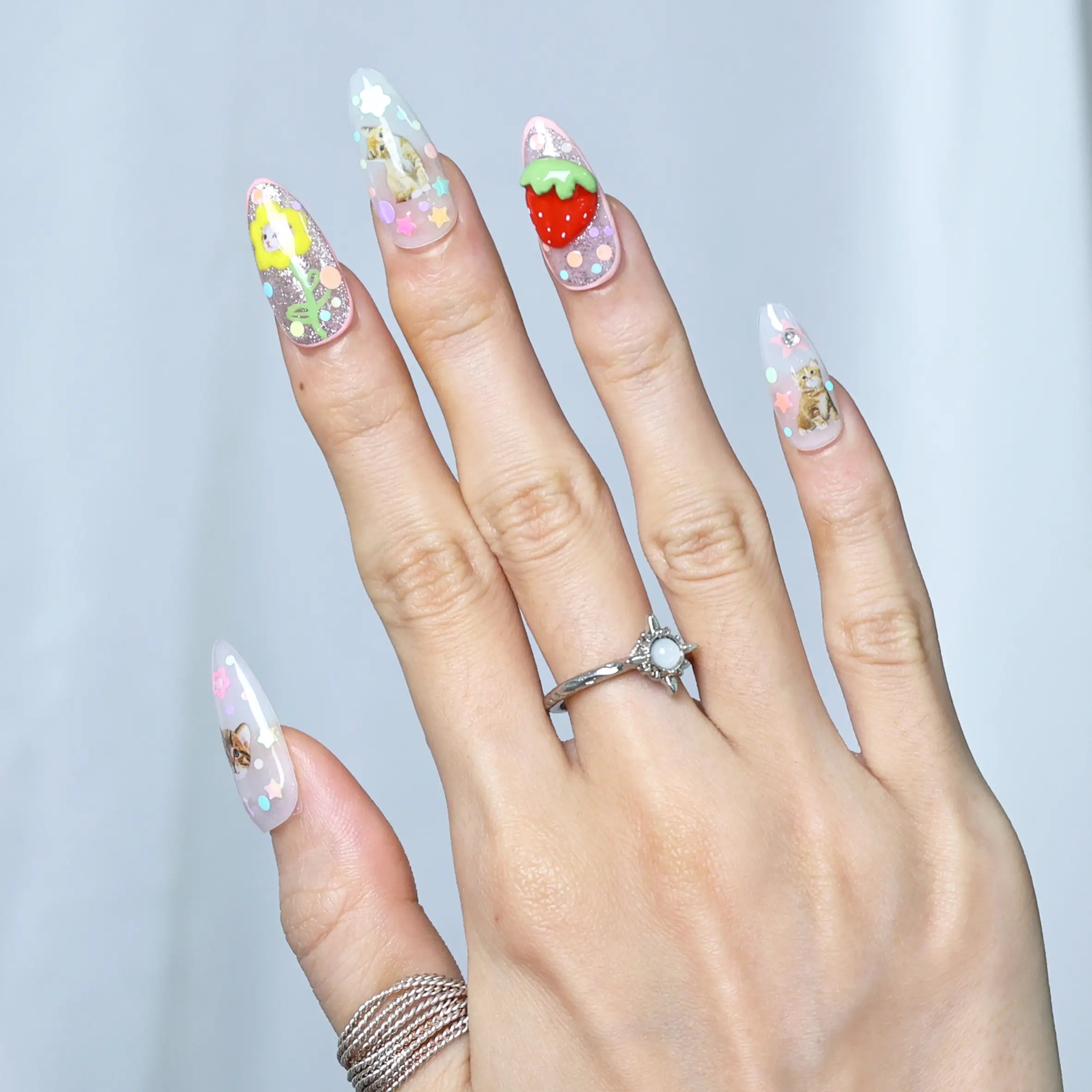 Hand pose highlighting vibrant pink nails with handcrafted kawaii figures, flower charms, and fruity sparkle