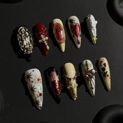 Long red and black press-on stiletto nails with gothic crosses, bleeding hearts, celestial gold stars, and religious sculptures.
