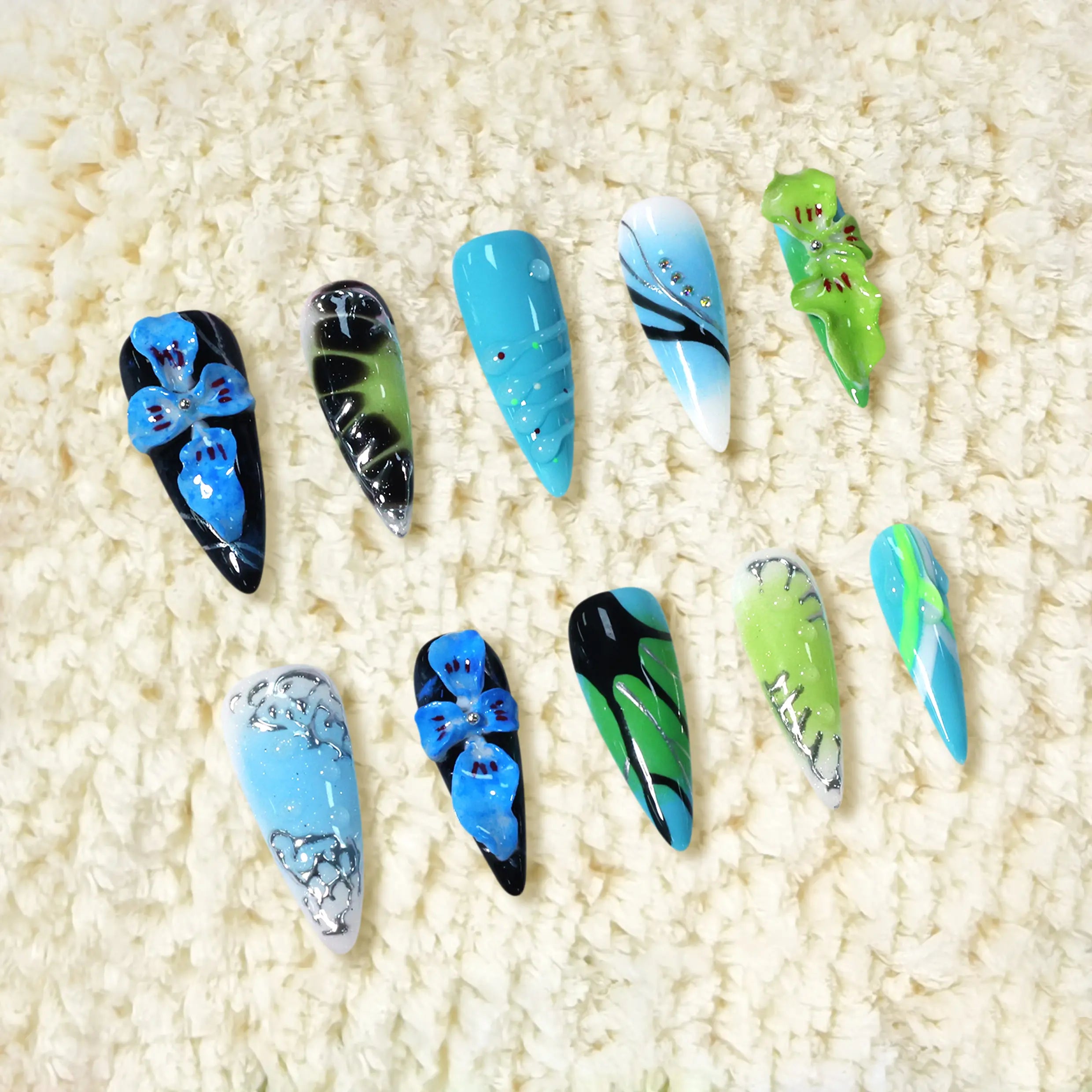 Vibrant long stiletto press-on nails with black, blue, and green tones, featuring 3D floral accents and rhinestone details.