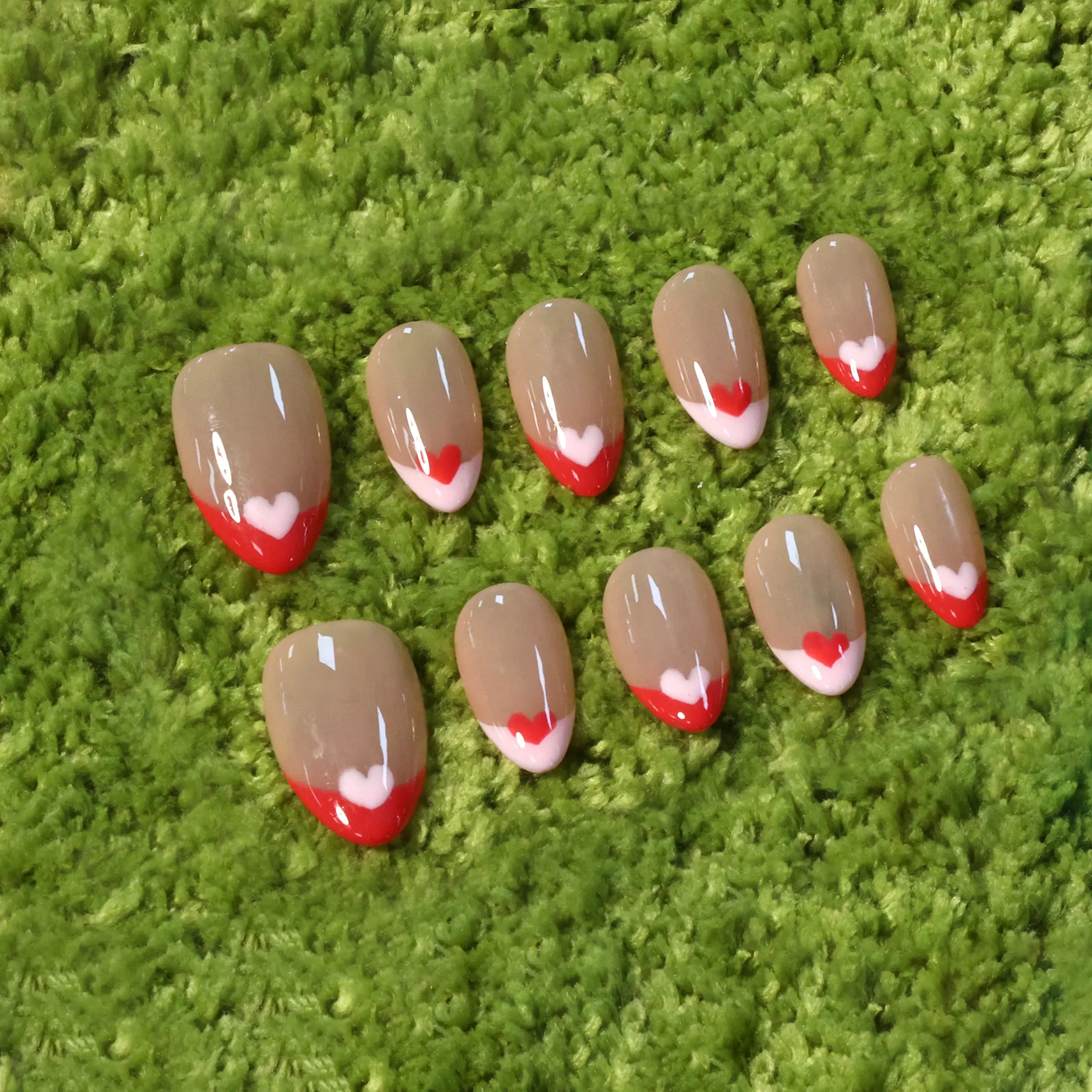 Short almond press-on nails with nude base, red French tips, and white heart accents for a romantic manicure.